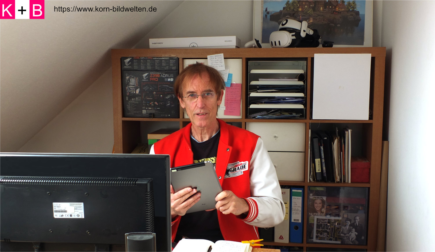 Andreas U. Korn im Buero am Arbeitsplatz mit iPad und roter Jacke.